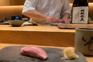 In the foreground, a piece of otoro nigiri with rich, marbling and a small mound of pickled ginger rest on a dark grey stone serving plate alongside a ceramic green tea cup. Behind the wooden counter, the blurred upper torso of a sushi chef in a white chef jacket works, positioned next to a sake bottle with Japanese calligraphy on its white label.
