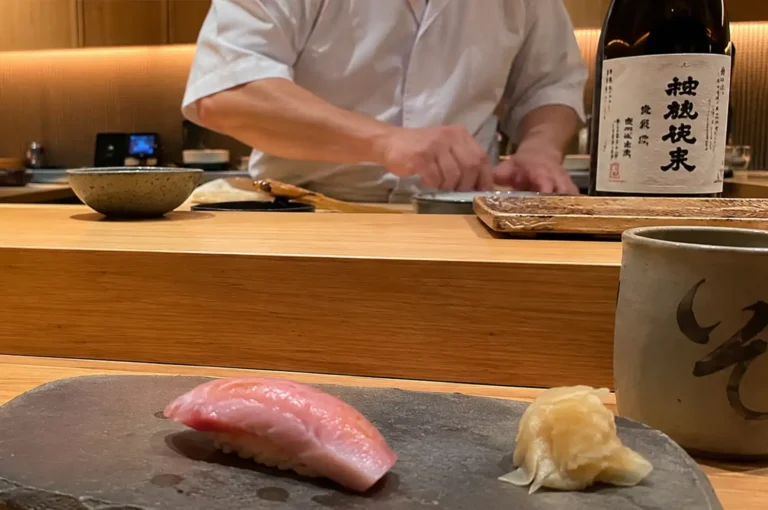 In the foreground, a piece of otoro nigiri with rich, marbling and a small mound of pickled ginger rest on a dark grey stone serving plate alongside a ceramic green tea cup. Behind the wooden counter, the blurred upper torso of a sushi chef in a white chef jacket works, positioned next to a sake bottle with Japanese calligraphy on its white label.