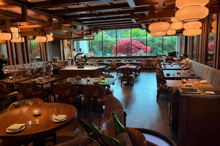 A wide view showcases the spacious interior of an upscale, modern Japanese restaurant featuring warm wooden architectural beams on the ceiling, atmospheric glowing lanterns, and dark wood tables paired with patterned armchairs. Large floor-to-ceiling glass windows at the back look out onto lush green foliage centered around vibrant red-leafed trees, with chefs visibly working behind a long sushi counter in the background.