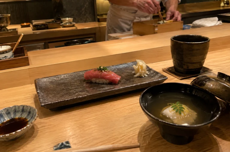 A diner’s perspective at an intimate omakase counter, featuring a long, rectangular dark ceramic plate holding a piece of tuna nigiri garnished with finely chopped scallions alongside pickled ginger. A black bowl filled with clear broth containing a delicate piece of fish topped with citrus zest rests on the right next to a rustic ceramic teacup, with a small soy sauce dish and chopsticks in the foreground. In the soft-focused background, a chef in a white uniform works behind the smooth wooden counter using wooden chopsticks to arrange dishes.
