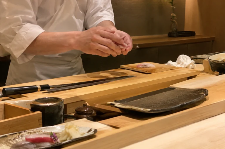 A low-angle, eye-level shot captures a sushi chef in a white uniform shaping nigiri with focused precision behind a light wooden omakase counter. On the wooden prep block sits a long, sleek yanagiba knife and a fresh slice of fish, while the foreground displays an earthy ceramic platter with pickled ginger and sashimi, a dark sauce cup, and a stone serving dish set against warm, warm-toned dining room lighting.