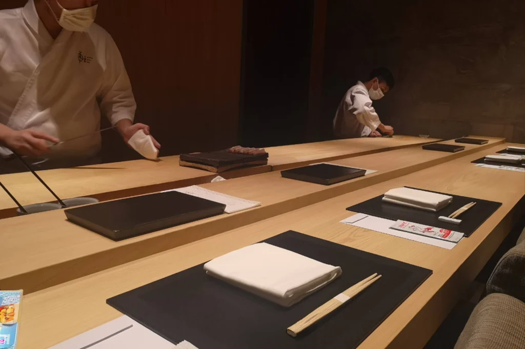 An interior view of an omakase dining counter set with minimalist place settings, each featuring a black mat, white folded napkin, and wooden chopsticks, while two chefs in white coats and masks work quietly in the background along the sleek wooden bar.