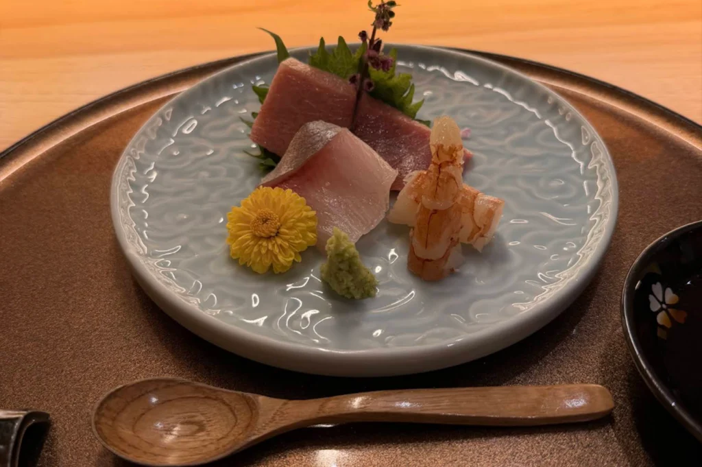 An eye-level shot of a refined sashimi course served on a light blue patterned ceramic plate, set upon a dark round tray. The dish features precise slices of fresh raw fish, a curled cooked prawn, a small dab of fresh wasabi, and a bright yellow edible flower garnish accompanied by a sprig of purple shiso blossom.