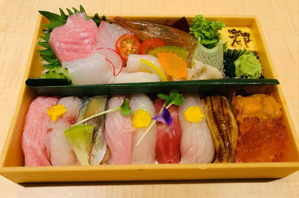 An overhead view of a premium bento box neatly arranged with fresh Japanese delicacies, divided into two distinct compartments. The top section contains vibrant sashimi slices including otoro, white fish, a sweet botanical prawn, cucumber, and a tamago slice stamped with Japanese characters, while the bottom section holds an array of nigiri sushi pieces, a glazed eel slice, and a side filled with glowing orange ikura and sea urchin.