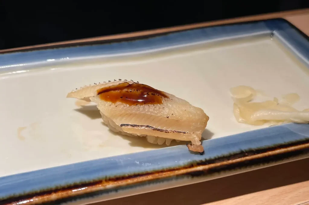 A single piece of aburi nigiri sushi features lightly torched white fish brushed with a glossy dark sweet soy glaze, presented on a light ceramic rectangular plate alongside pickled ginger slices.