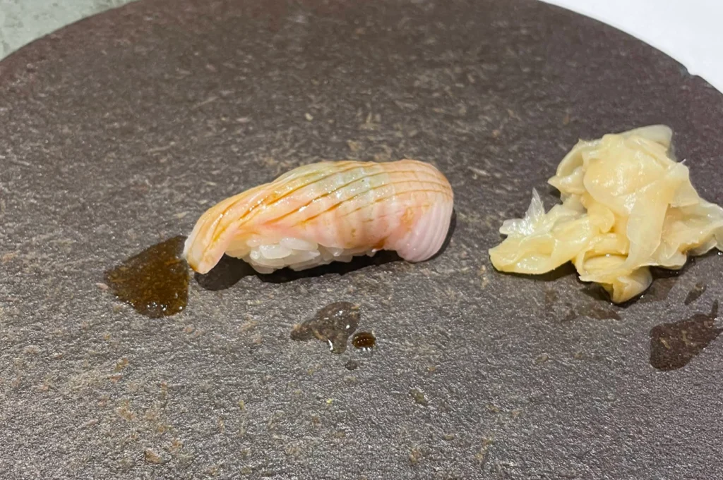 A single piece of nigiri sushi brushed with a glistening coat of soy sauce sits on a textured dark gray stone plate next to a mound of pale pickled ginger slices. The delicate slice of fish rests over seasoned sushi rice, leaving a slight soy sauce residue on the stone surface.