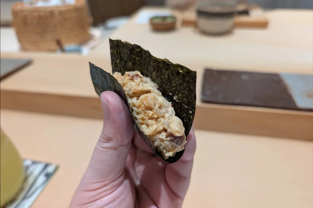 A person holds a hand roll wrapped in a crisp, dark green sheet of nori seaweed, exposing a generous filling of creamy chopped fish mixed with seasoned sushi rice. In the softly blurred background, a light wooden omakase counter displays small ceramic dishware, a wooden tray, and a woven basket.
