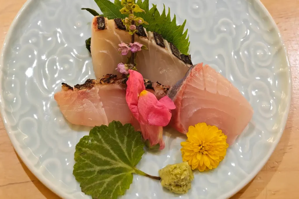 A light blue ceramic plate with embossed wave patterns showcases three fresh sashim cuts, including seared white fish with charred skin and raw pink fish. The arrangement is delicately garnished with a bright yellow chrysanthemum, a pink edible flower, green shiso leaves, and a dollop of fresh wasabi paste.