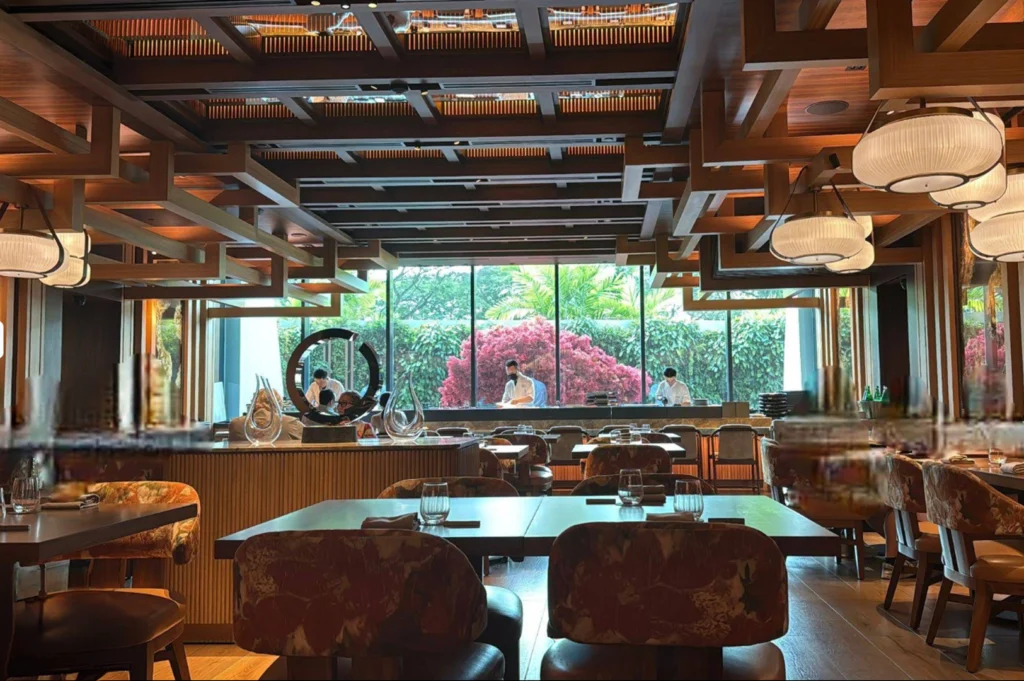 An eye-level shot captures a dining room layout set with dark wooden tables, patterned upholstered chairs, and warm paper lantern lights hanging from a decorative grid ceiling. Framed through the glass backdrop in the background is a open counter kitchen where staff in white uniforms prepare food against a garden view with bright pink foliage.