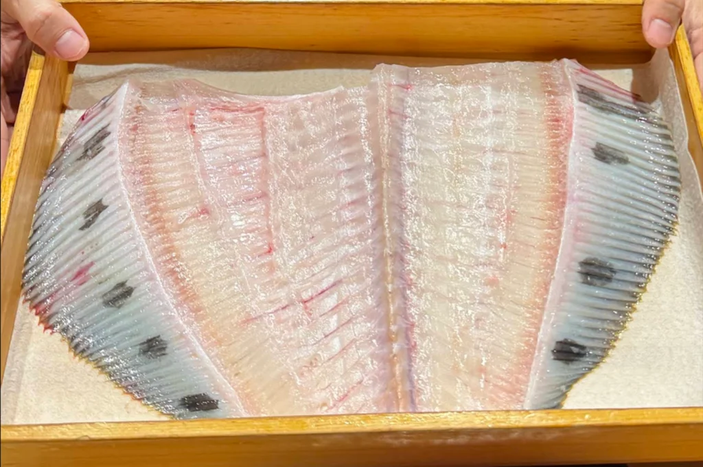 A close-up view looking down into a wooden presentation box held by a chef, displaying a large, butterfly-cut side of raw flatfish with intact dark fin edges and delicate white flesh arranged on parchment paper.