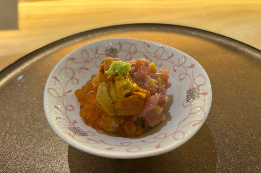 A close-up view of a delicate white porcelain dish decorated with faint pink line patterns, holding a mini chirashi bowl course. The dish is layered with seasoned sushi rice, glistening orange salmon roe (ikura), chopped fatty tuna (negitoro), bright yellow sea urchin (uni), and a small dollop of wasabi on top.