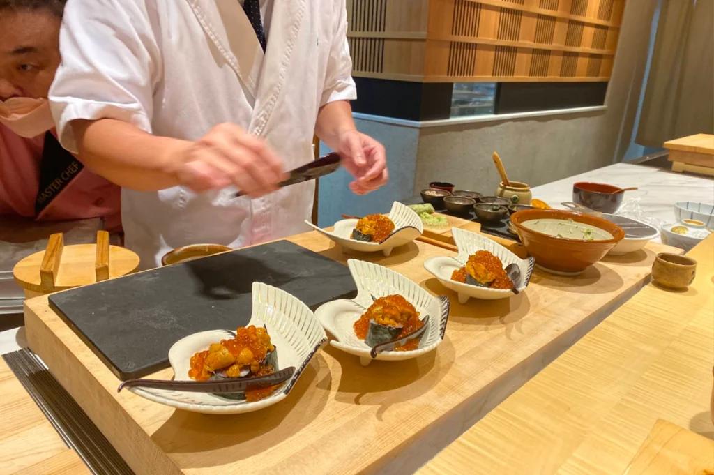 A chef clad in a white uniform assembly-lining specialty sushi courses along a wooden counter, carefully positioning ingredients over rowed dishes. Four individual shell-shaped white ceramic serving plates hold gunkan sushi topped generously with bright sea urchin (uni) and glistening salmon roe (ikura).