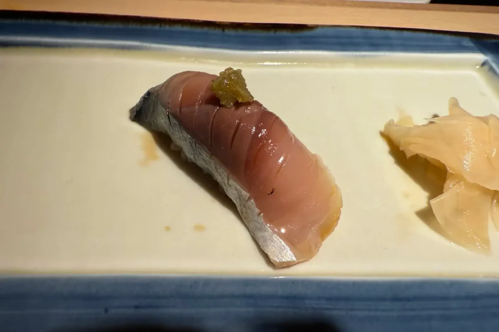 A close-up shot of a piece of mackerel nigiri sushi garnished with a dab of citrusy condiment on top, served on a light rectangular ceramic plate with a cluster of thinly sliced pickled ginger on the right side.