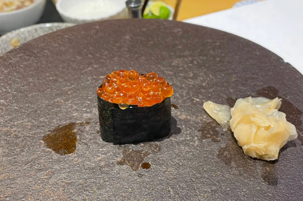 A single gunkan maki sushi piece wrapped in dark nori seaweed is piled high with glossy, translucent orange ikura (salmon roe) that catches the light. It is presented on a textured, dark round stone serving plate beside a small pile of pickled ginger.