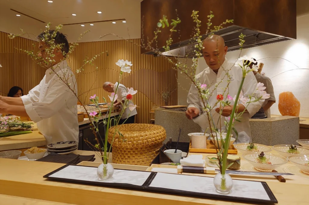 Behind a light wood omakase counter, sushi chefs in white coats meticulously assemble dishes framed by delicate floral arrangements in glass vases. Small clear bowls of food and dining utensils line the foreground tray, while warm ambient lighting, vertical wood-paneled walls, a large metal extraction hood, and a block of Himalayan pink salt set the serene, high-end atmosphere.
