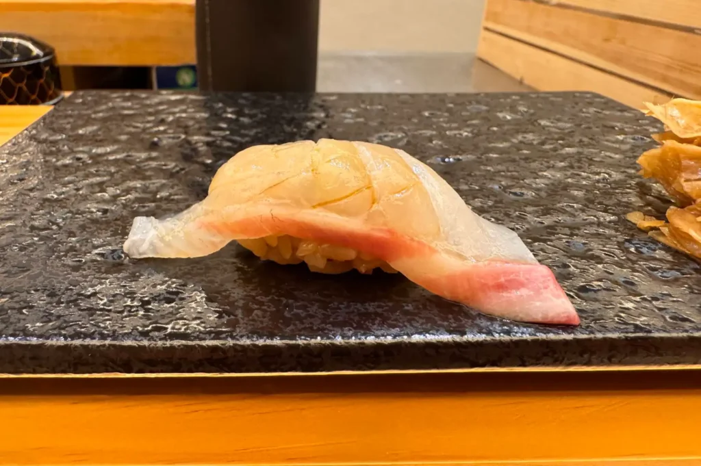 A single piece of tai nigiri featuring a delicate, translucent slice of white fish with pink edges over seasoned sushi rice rests centered on a reflective black slate tray. To the right edge of the tray, a small cluster of thinly sliced pickled ginger sits on the dark, textured surface.