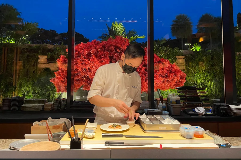 A chef wearing a white uniform and black face mask attentively garnishes a plate of thinly sliced carpaccio at a clean, light-wood sushi prep station inside a Japanese restaurant. In the background behind glass walls, bright red autumn foliage stands out against a deep blue evening dusk light.