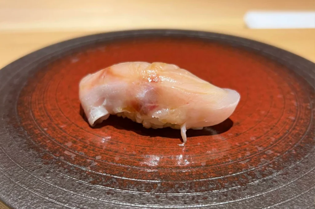 A piece of sea bass nigiri with translucent, lightly glazed white fish draped over sushi rice, presented in the center of a rustic, round ceramic plate with textured concentric circles and earthy red-brown hues.
