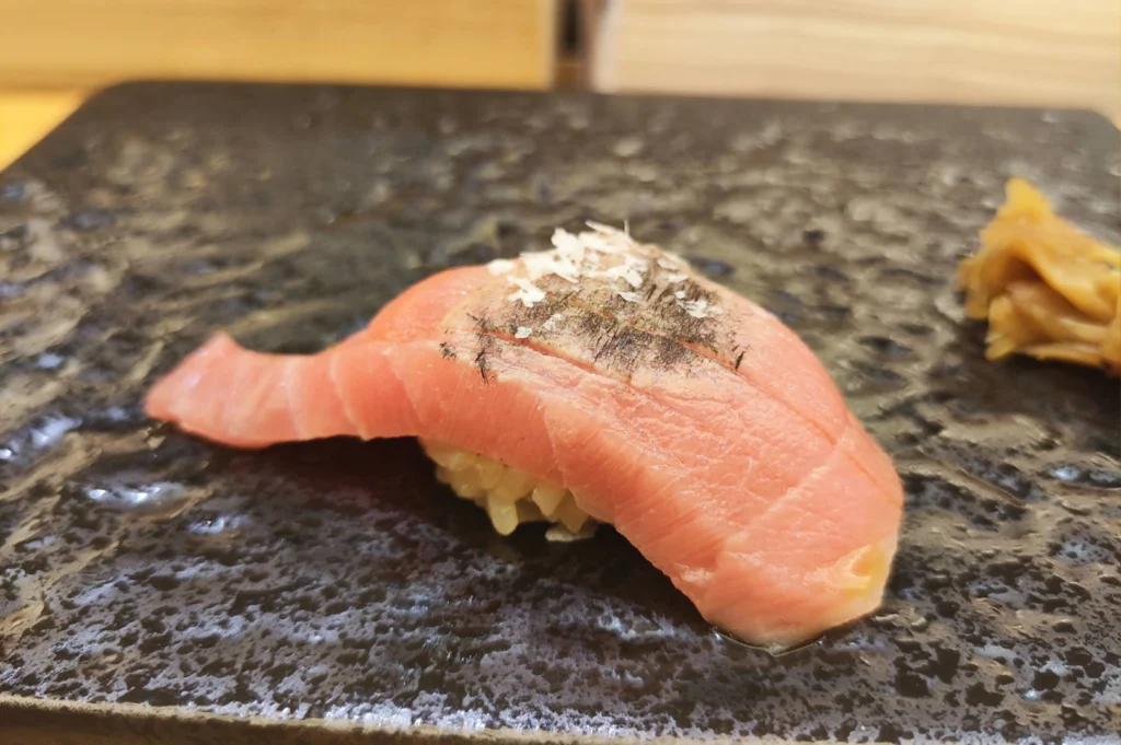 A close-up shot of a single piece of nigiri sushi resting on a dark, textured slate serving plate. The piece features a tender slice of lightly seared pink otoro draped over seasoned sushi rice, finished with a subtle sear mark, sea salt flakes, and pickled ginger placed to the side.