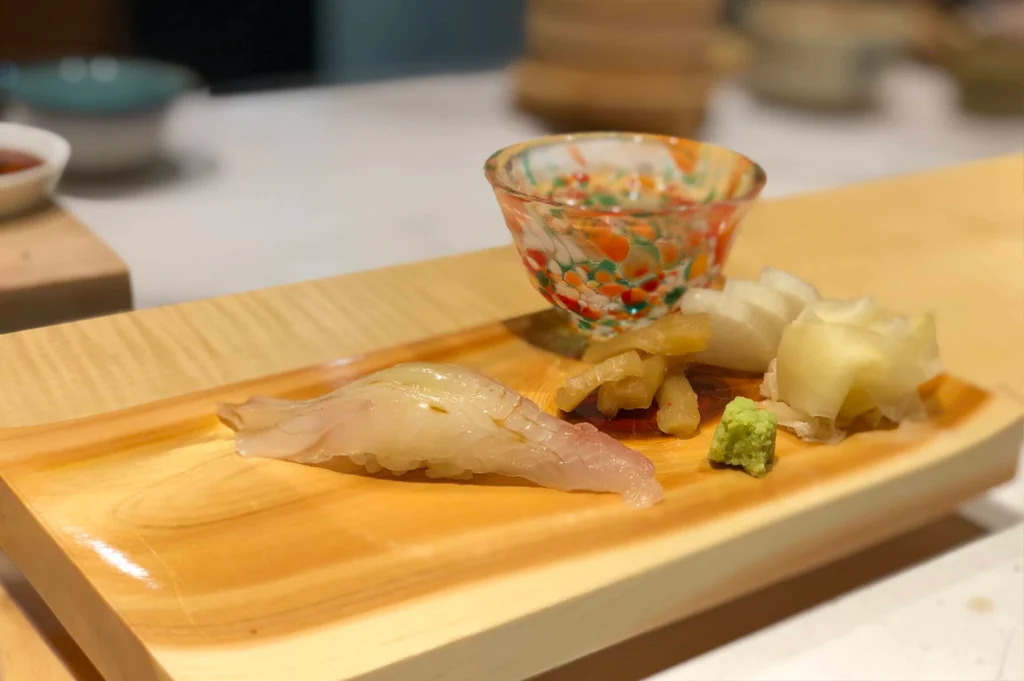 A close-up shot of a delicate piece of white-fish nigiri served on a curved wooden sushi board, brushed with a light glaze. Accompanied in the background by a small colorful terrazzo glass cup, pickled ginger slices, pickled radish, and a dollop of fresh green wasabi.
