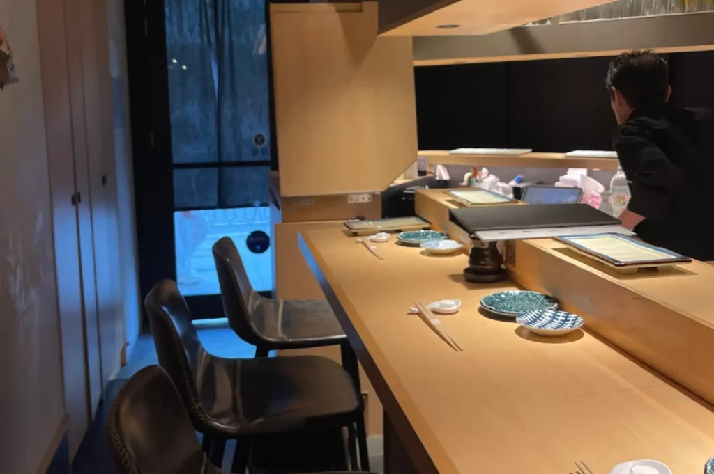 An interior view of an intimate, modern sushi bar showing a light wood counter set with small ceramic dishes and chopsticks, high-backed leather dining chairs lined along the seating area, and a chef working behind the counter space.