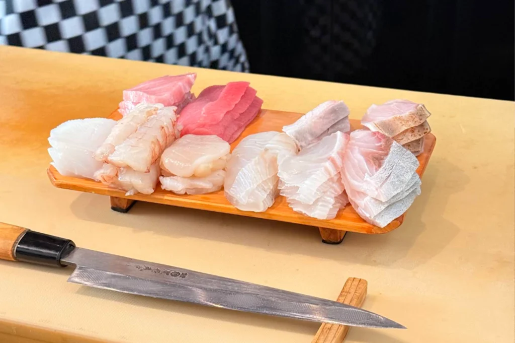 A wooden platter set on a bright wooden counter displays an arrangement of neatly sliced raw fish and seafood ingredients prepared for omakase service, including bright red tuna, pale white fish, scallops, and sweet prawns. In the foreground, a long, sharp Japanese yanagiba knife rests flat on a wooden block next to the platter.