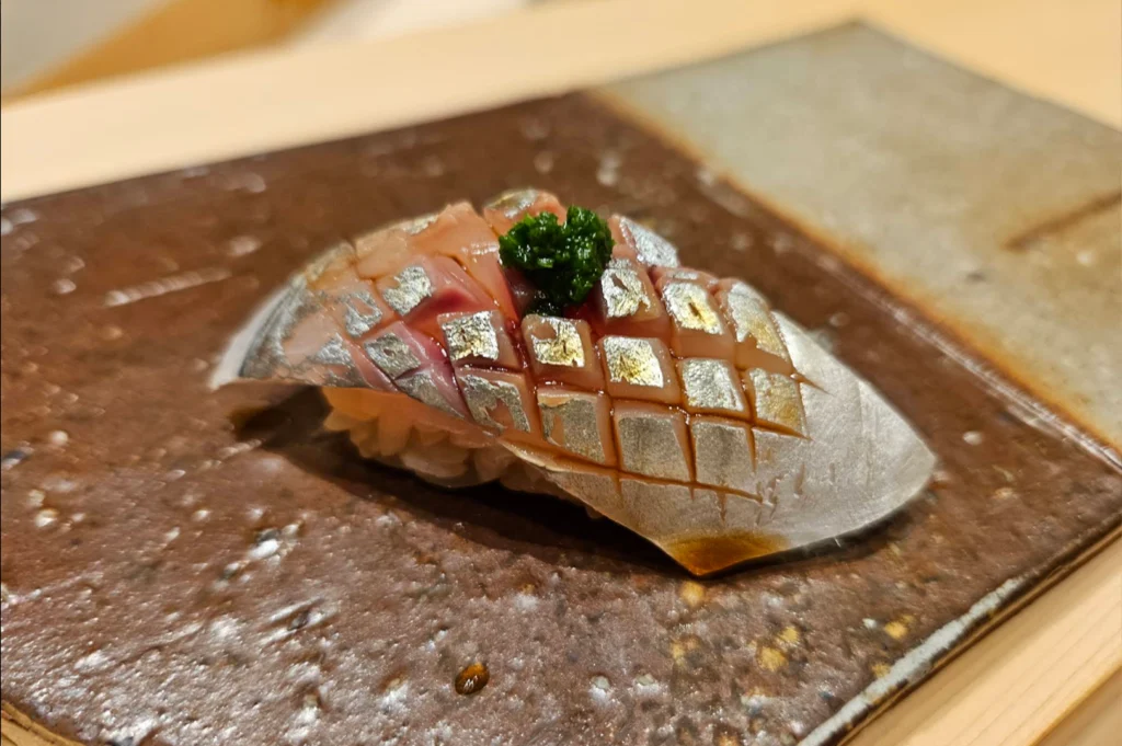 A single piece of nigiri sushi sits centered on a dark, textured rectangular ceramic plate. The slice of fish features shiny silver skin with a precise cross-hatched scoring pattern, brushed lightly with a glint of soy glaze and topped with a small dollop of green finely chopped scallion or herb paste.