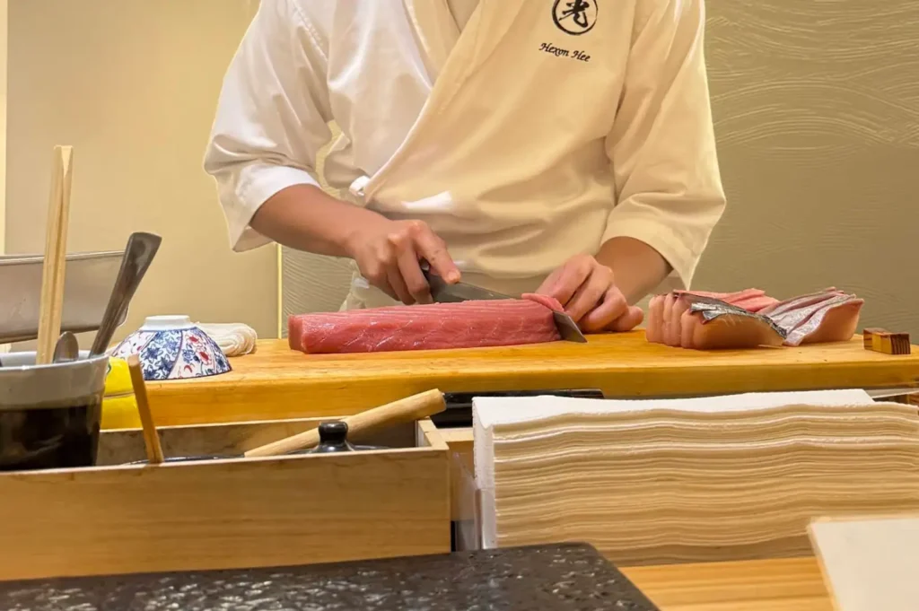 A sushi chef wearing a white jacket embroidered with "Hexon Hee" cuts a thick, vibrant red saku block of tuna on a wooden prep counter. To the right of the tuna loin rest several freshly sliced cuts of sashim—including oily fish with silver skin—while utensils, small ceramic dipping bowls, and stacked white paper towels occupy the foreground.