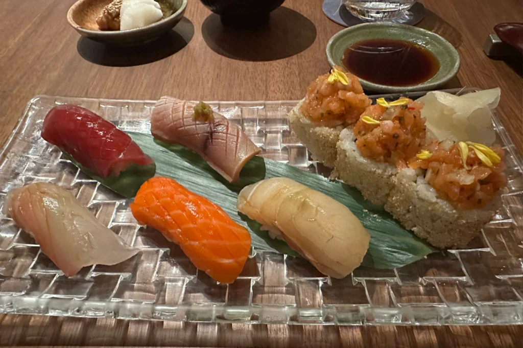 A close-up shot presents an assortment of fresh sushi arranged on a clear, textured glass rectangular platter lined with a green leaf. The platter holds five pieces of nigiri—including tuna, salmon, and white fish—alongside three crisp-coated maki rolls topped with minced fish, accompanied by pickled ginger and a small dish of soy sauce.