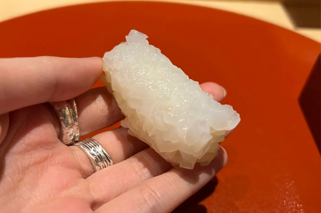 A hand wearing silver rings holds a single piece of squid nigiri with intricately scored, translucent white squid glistening over sushi rice, shown close up against a warm, minimalist background.