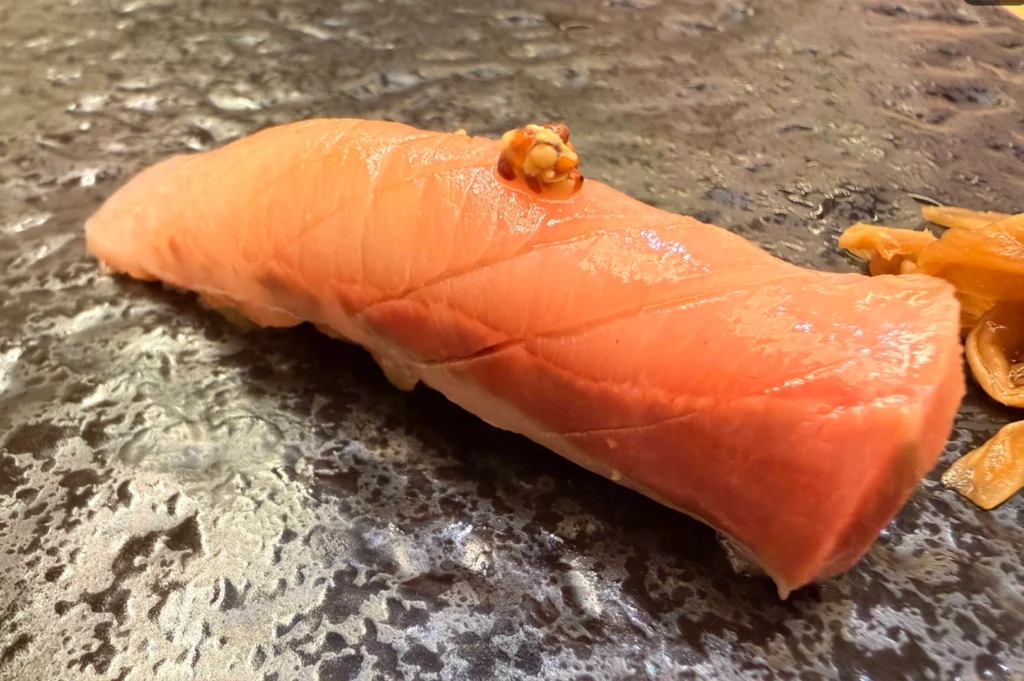 A close-up, angled view of a single piece of tuna nigiri presented on a dark, glossy stone platter. The thick, vibrant pink slice of fish has precise scoring marks along its length and is delicately topped with a small speck of relish or condiment in the center.