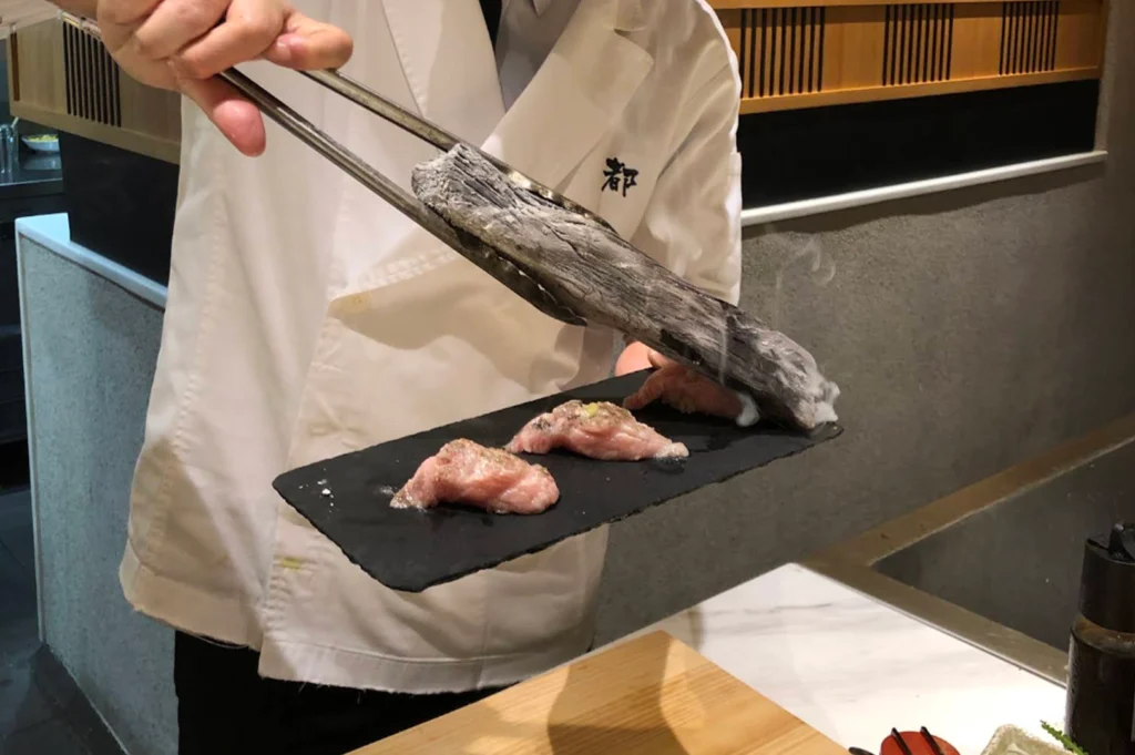 A close-up action shot of a chef using metal tongs to press a piece of glowing hot binchotan charcoal directly onto slices of marbled nigiri sushi placed on a black slate platter, creating a gentle wisp of smoke to sear the fish.