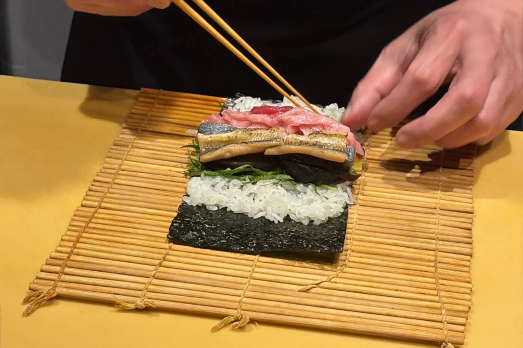 A chef's hands use wooden chopsticks to assemble a futomaki roll over a sheet of nori seaweed and sushi rice laid out on a traditional bamboo rolling mat. The rich filling layers include strips of unagi (eel), tuna, shiso leaves, and pickled vegetables.