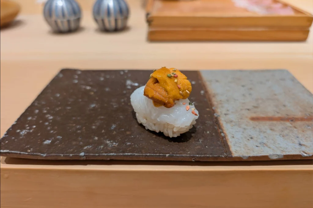 A single piece of nigiri sushi topped with translucent raw squid and a dollop of bright orange sea urchin (uni) rests on a dark ceramic platter with a gray glazed edge. The uni is sprinkled with colorful sesame seeds and furikake seasoning, with small ceramic cups and wooden serving trays blurred in the background on the light wooden counter.