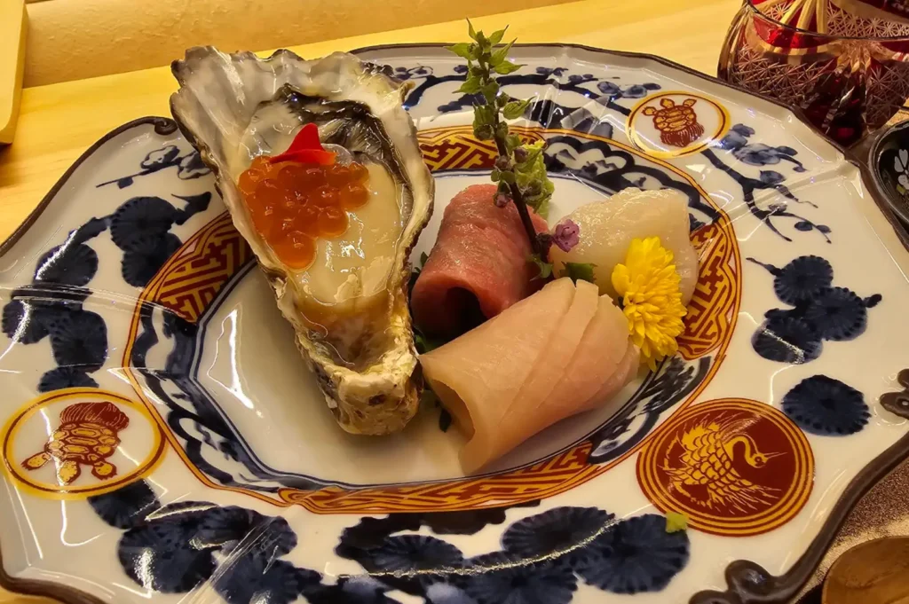 An artistic seafood course is served on an ornate, hexagonal ceramic plate decorated with traditional blue and yellow motifs. The dish presents a plump, fresh oyster on the half shell glistening with sauce and topped with shiny salmon roe, surrounded by neatly rolled slices of raw fish, a scallop, a yellow chrysanthemum flower, and a delicate sprig of shiso blossoms.