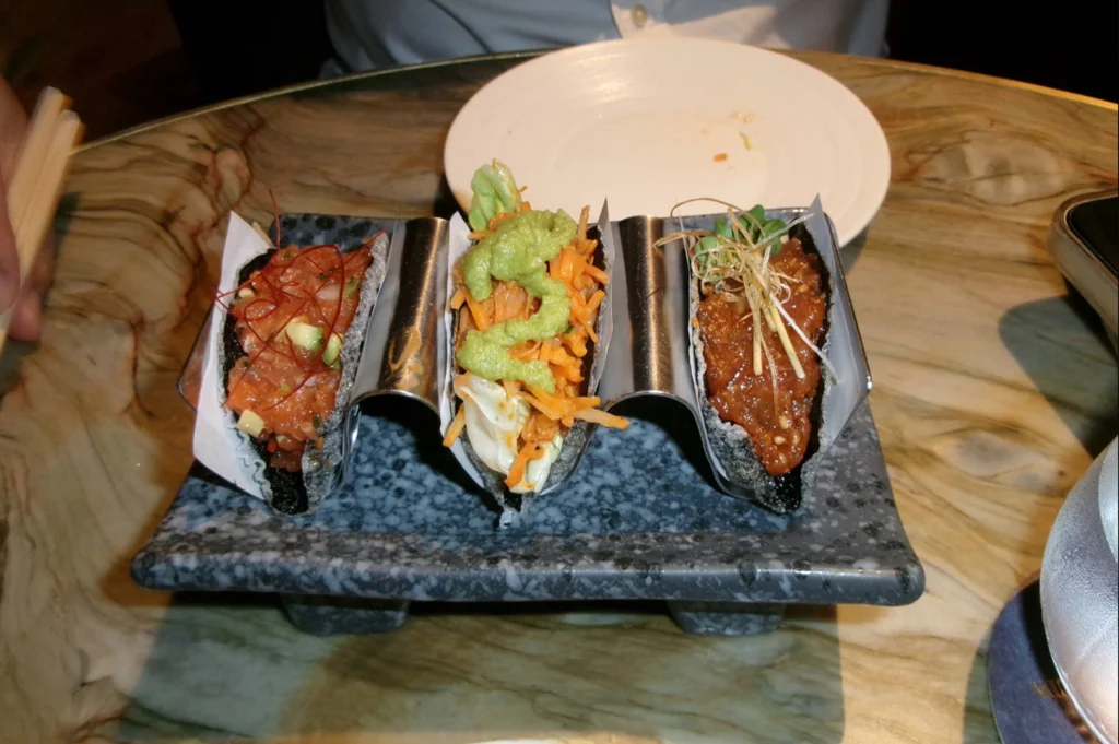 A top-down shot features three crispy nori taco-style bites held upright in a metal holder resting on a gray stone plate over a marble tabletop. Each taco shell is filled with different toppings, including minced red tuna, shredded vegetables with a dollop of green guacamole, and a savory seasoned meat mixture garnished with crispy strands.