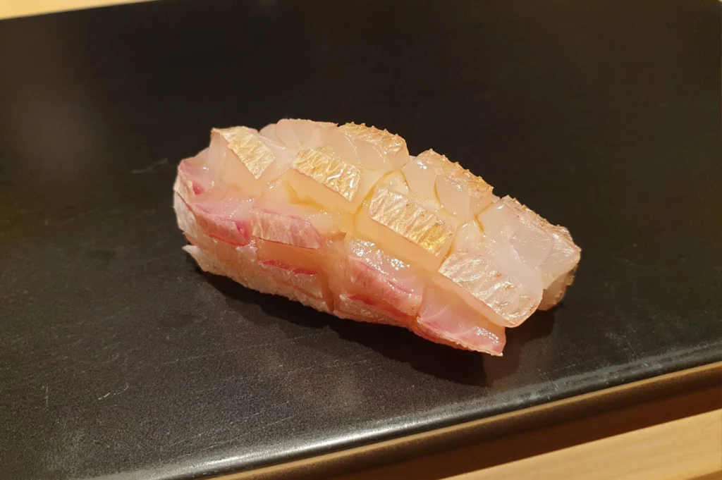 A close-up of a piece of sea bream nigiri resting on a smooth black slate platter, featuring lightly torched, cross-hatched skin over delicate pink-hued fish and seasoned sushi rice.