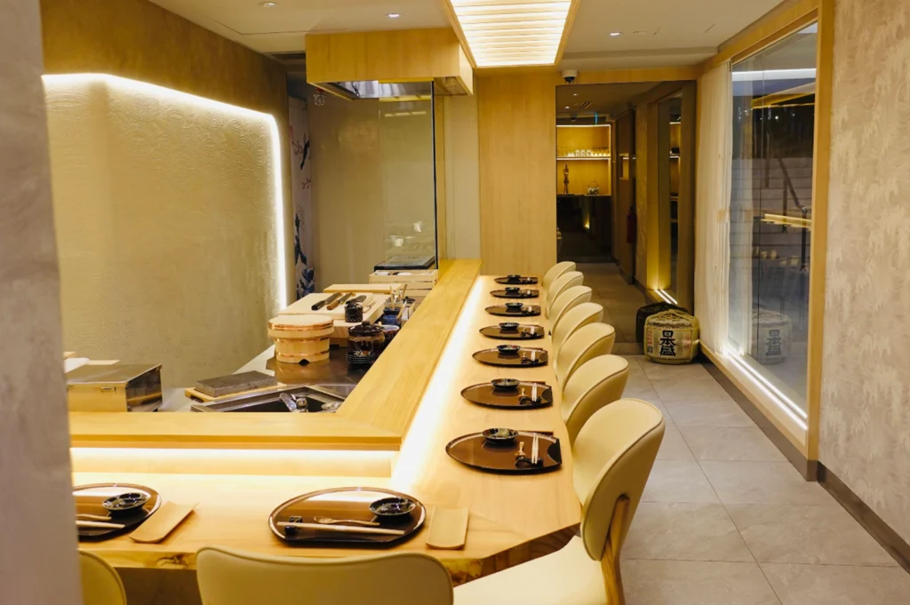 A wide interior view of an elegant, modern omakase sushi restaurant featuring light wood finishes and warm ambient lighting. A long, L-shaped wooden sushi counter lined with sleek beige armchairs spans the room, with individual place settings arranged neatly along the counter, while traditional sake barrels rest against a textured accent wall in the background.