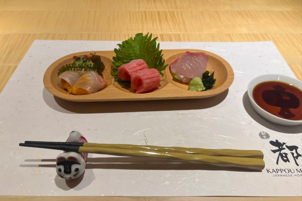 An aesthetic place setting on a white paper placemat featuring a wooden three-compartment dish holding fresh sashimi offerings, including sliced tuna rolls, white fish, and sea urchin wrapped in fish. Next to the dish sit light-green wooden chopsticks resting on a small ceramic cat rest and a small dish of soy sauce.