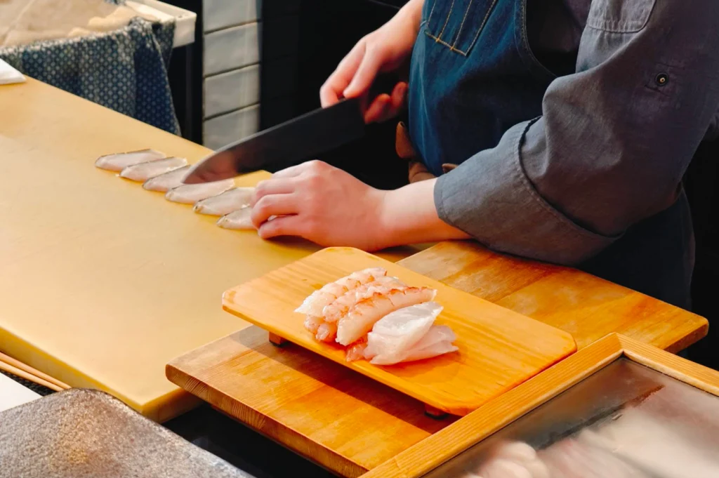 A sushi chef wearing a dark blue apron uses a sharp sashimi knife to carefully slice fresh fish on a light-colored wooden prep counter. Next to the cutting area, a small wooden pedestal holds a fresh stack of cooked sweet prawns and pale white fish fillets ready for plating.