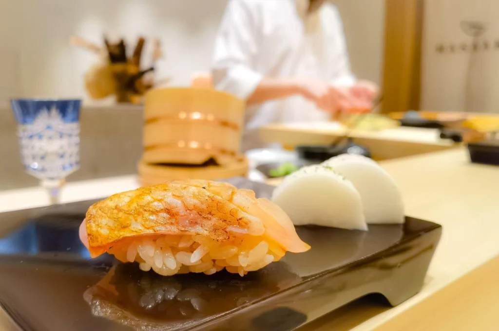 A close-up shot on a glossy black ceramic serving block shows a piece of seared nigiri sushi with golden, lightly charred fish skin alongside two thick slices of pickled white radish. In the soft-focus background, a chef in a white uniform works at the counter next to a traditional wooden rice tub and a cut-crystal glass.