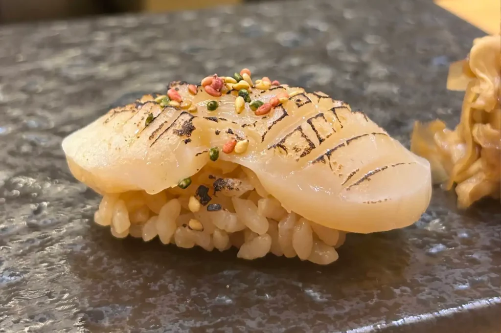 A piece of seared scallop nigiri sits on a dark, textured stone platter next to a small pile of pickled ginger. The scallop slice is lightly seared with crosshatch score marks along its top surface and sprinkled with colourful toasted sesame seeds and furikake seasoning over season rice.