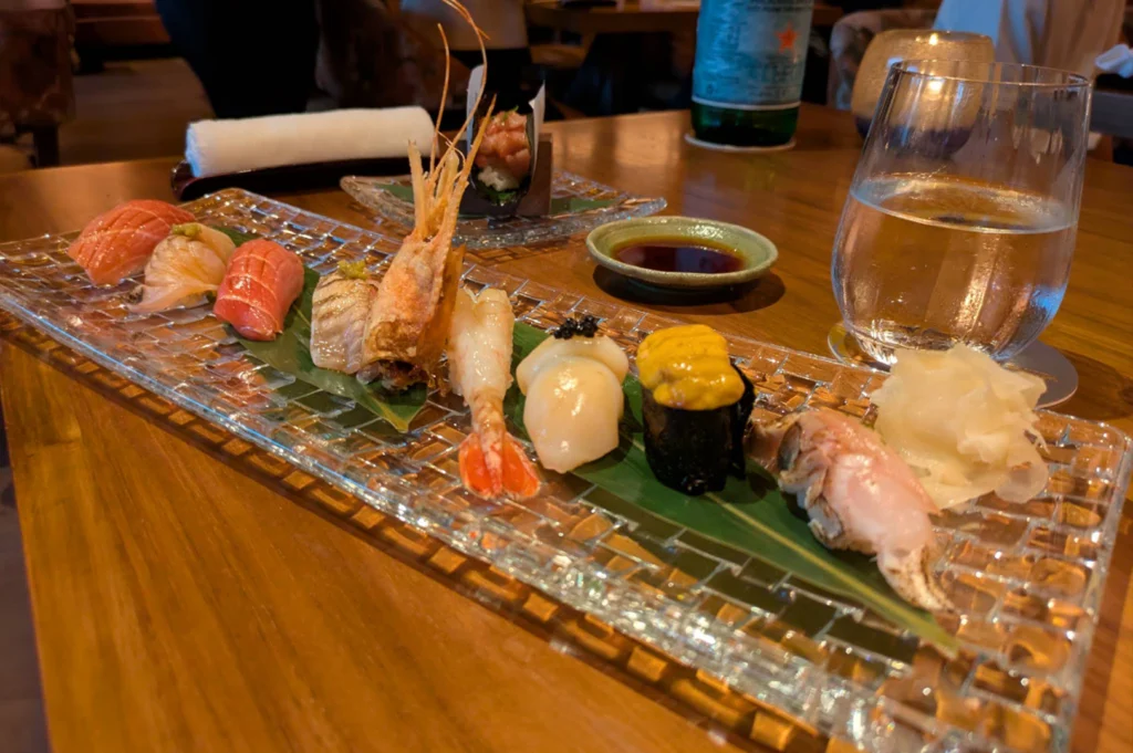 A long rectangular glass platter resting on a warm wooden dining table showcases a premium assortment of nigiri sushi, featuring fresh cuts of salmon, tuna, sweet botanical shrimp with head attached, scallop topped with caviar, sea urchin (uni) gunkan, and crab leg. Next to the platter sit pickled ginger, a small dish of soy sauce, a glass of water, and a rolled hot towel.