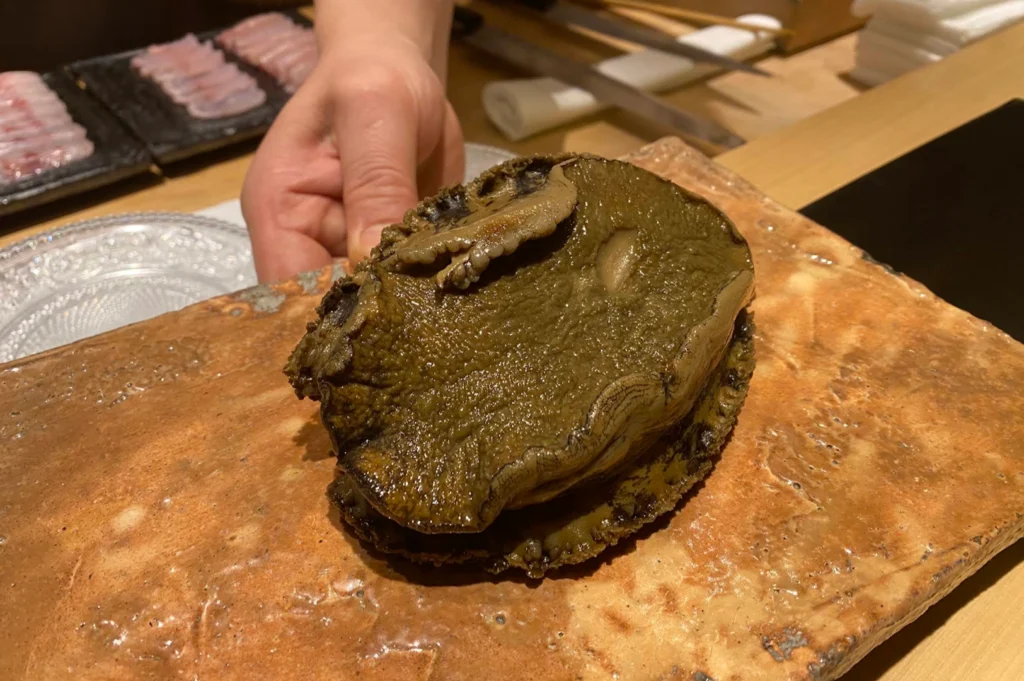 A chef holds up a large, textured whole abalone presented on a rustic, rectangular stoneware plate, with pre-sliced raw fish portions visible on a black dish in the blurred background.