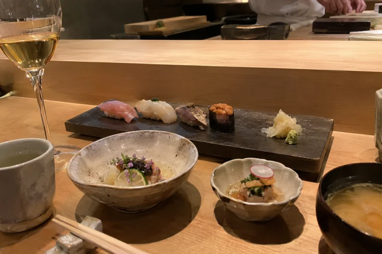 A warm, eye-level shot captures an intimate omakase dining experience laid out across a smooth light-wood counter. In the mid-ground, a long, dark slate platter holds four delicate pieces of sushi—including fresh nigiri topped with tuna, white fish, seared fish, and bright orange uni gunkan—alongside pickled ginger and wasabi. Arranged in the foreground are various ceramic dishes containing delicate appetizers decorated with edible flowers and citrus slices, accompanied by a small bowl of miso soup, a cup of green tea, wooden chopsticks on a rest, and a filled wine glass standing to the left. In the soft-focus background, the hands of a sushi chef in a white uniform can be seen preparing dishes behind the wooden bar counter.