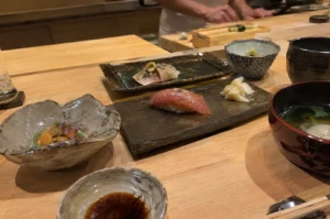 An intimate, high-angle view of an omakase meal laid out across a light-wood sushi counter, featuring a central dark ceramic platter holding a piece of glistening tuna nigiri sushi next to a small portion of pickled ginger (gari). Surrounding the main platter are various artisan ceramic dishes containing fresh sea urchin (uni) garnished with purple shiso blossoms, a secondary plated piece of dressed fish, a small blue-and-white bowl, a cup of dark broth, and a soy sauce dipping dish, with chopsticks resting nearby. In the softly blurred background, a sushi chef dressed in a white uniform works attentively behind the wooden counter prep area.