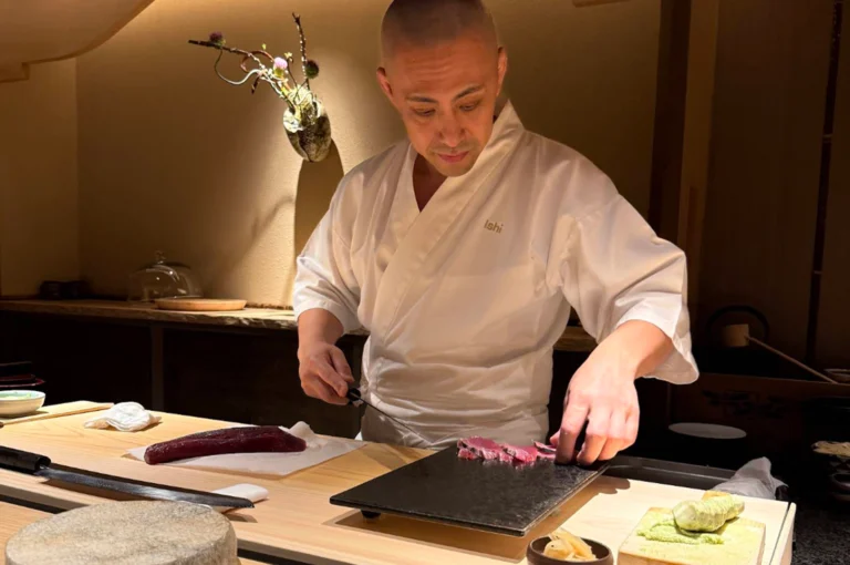 A focused Japanese sushi chef wearing a clean white chef coat with "Ishi" embroidered on the chest stands behind a light-colored wooden counter, carefully arranging freshly sliced, vibrant pink fish onto a dark, textured slate platter. The clean workstation includes a long, sharp sushi knife resting on the wooden counter, a block of fresh wasabi on a wooden grater, a small dish of pickled ginger, and a delicate wall-mounted floral arrangement casting warm light in the soft background.