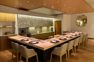 An wide interior perspective showcases an L-shaped light-wood omakase dining counter lined with comfortable tan upholstered armchairs set against wooden flooring. The minimalist Japanese interior features warm recessed ceiling lights, geometric wall tile accents, light wood paneling, and an abstract golden decorative mirror mounted on the far wall.