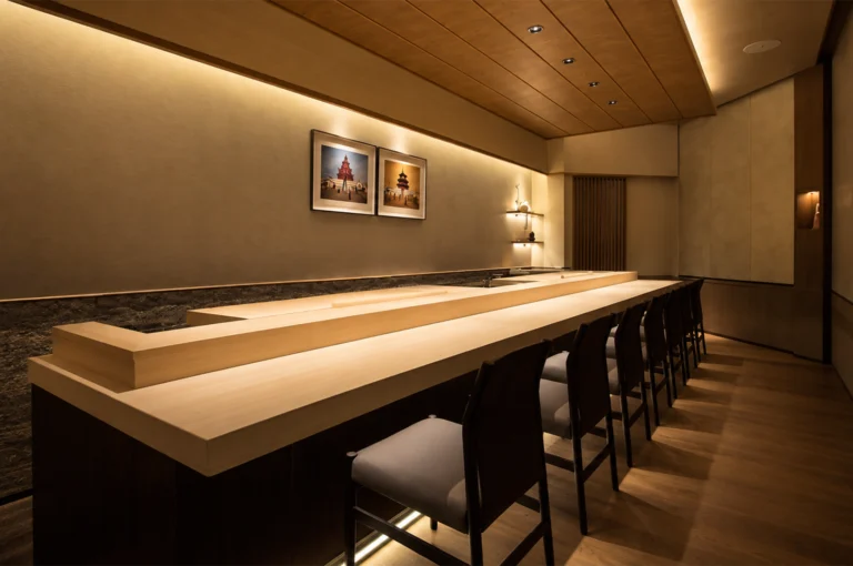 An interior view of an elegant, minimalist sushi restaurant featuring a long, polished light-wood dining counter lined with sleek black bar chairs with grey cushioned seats. Warm ambient lighting glows overhead and under the counter edges, illuminating neutral-toned walls, framed artwork depicting traditional Asian architecture, and floating wooden display shelves in the corner.