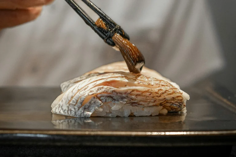 A macro, close-up shot captures a chef applying a light layer of glaze to a piece of seared fish nigiri using a small basting brush with dark wooden handles. The piece of sushi features lightly torched, textured fish skin resting atop seasoned rice, presented on a dark, glossy ceramic plate under direct dining lights.